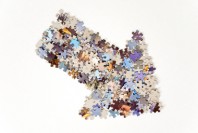 Thick arrow pointing to the left and up, made from plenty of colorful jigsaw puzzle pieces separated on white background.